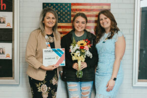 Three women stand together against a wall, smiling and looking at the camera, with one holding flowers in the center and one holding a certificate on the left.